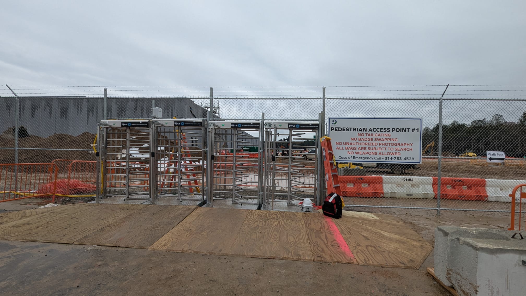 Full-height turnstiles placed at an entrance to a facility, alongside a fence with a barbed wire on top. 